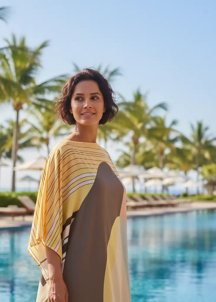 Woman in Vicharan HORIZON Yellow caftan standing by a pool with palm trees in the background at resort