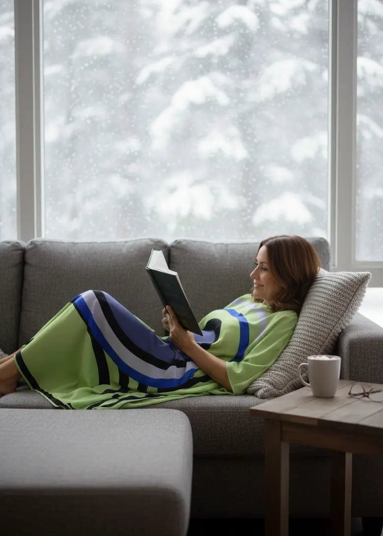 Woman in Vicharan MATCHA Green caftan reading a book on a couch with a snowy landscape outside the window